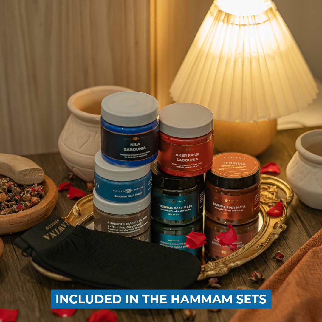 Zawina Morocco Hammam Sets product collection including Sabounia soaps, body masks, Rhassoul clay mask, and a black Kessa glove arranged on a gold tray with rose petals