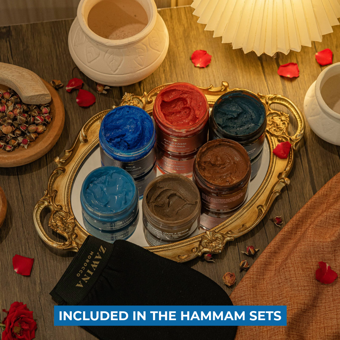 Zawina Morocco Hammam Sets product collection including Sabounia soaps, body masks, Rhassoul clay mask, and a black Kessa glove arranged on a gold tray with rose petals