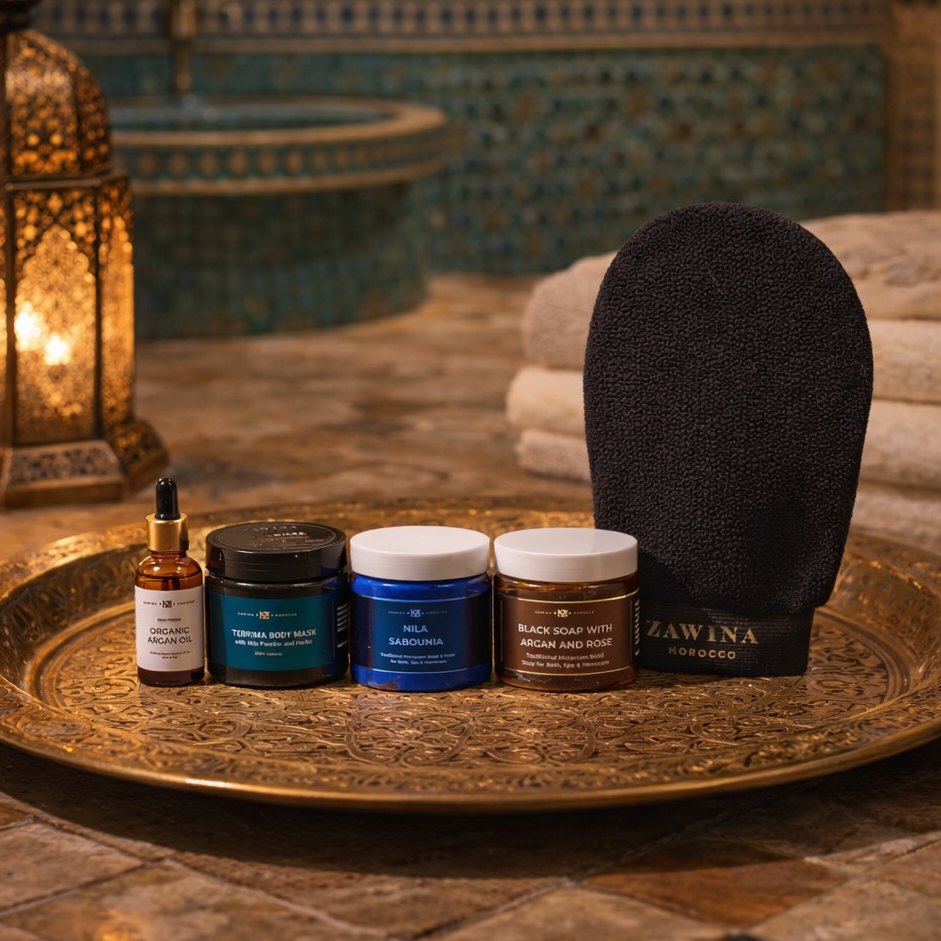 Zawina Morocco Nila Hammam Set featuring Organic Argan Oil, Tebrima Body Mask, Nila Sabounia, Black Soap with Argan and Rose, and a Kessa glove arranged on an engraved brass tray in a traditional hammam setting